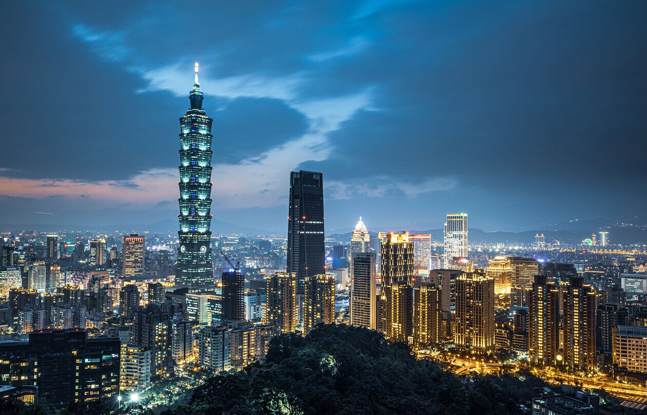 台北优化版 300 A panoramic view of a city skyline at night, featuring a tall, illuminated tower amidst various buildings and a cloudy sky.