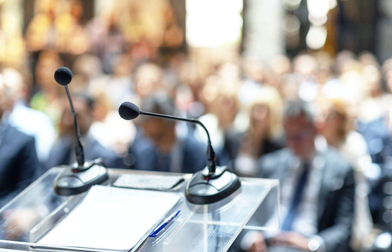 present_use_of_capsules_optimized_300 A podium with two microphones in focus, overlooking a blurred audience at a conference or event.