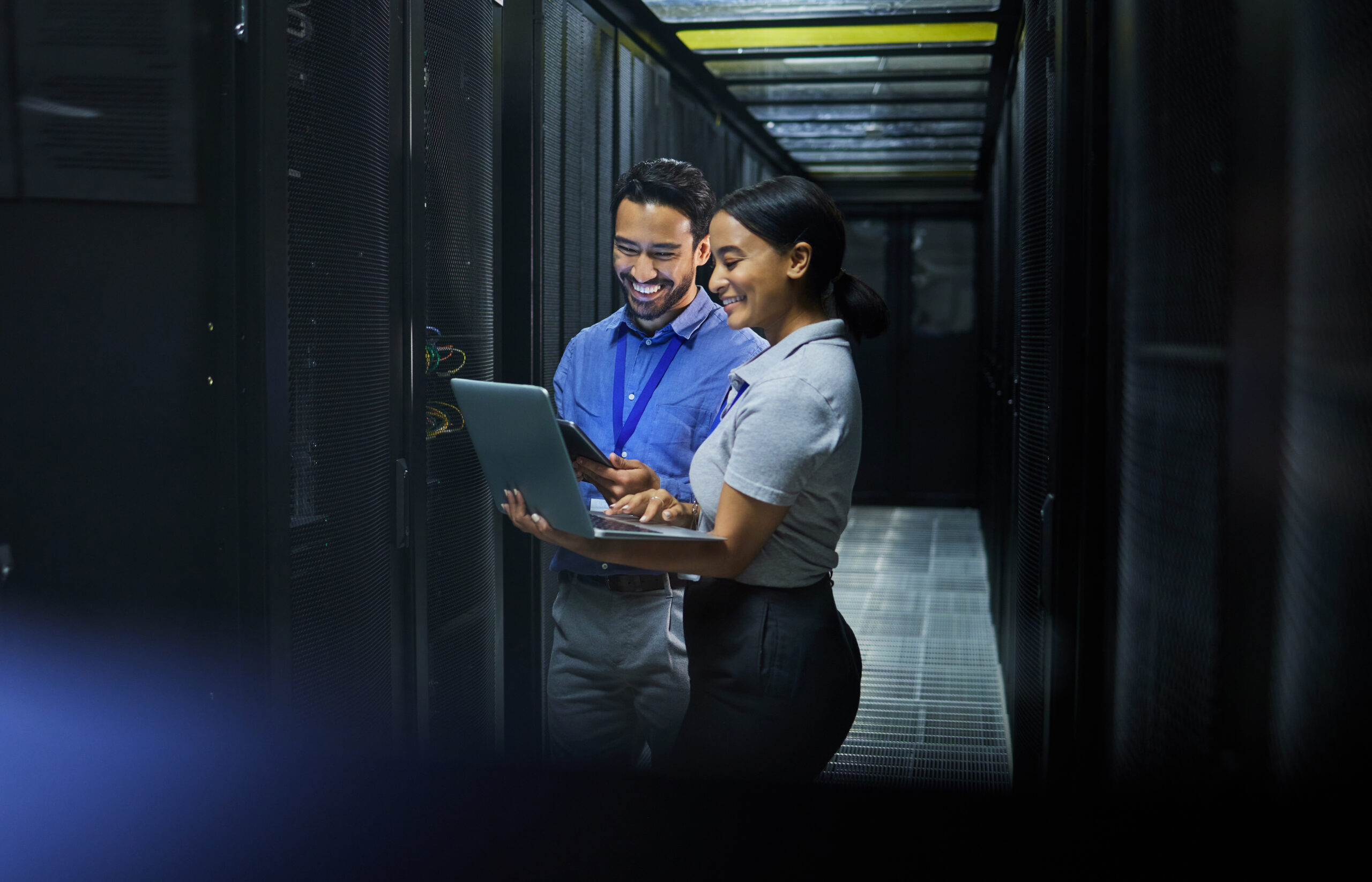 Two professionals are smiling and collaborating in a server room, one using a laptop and the other holding a tablet.