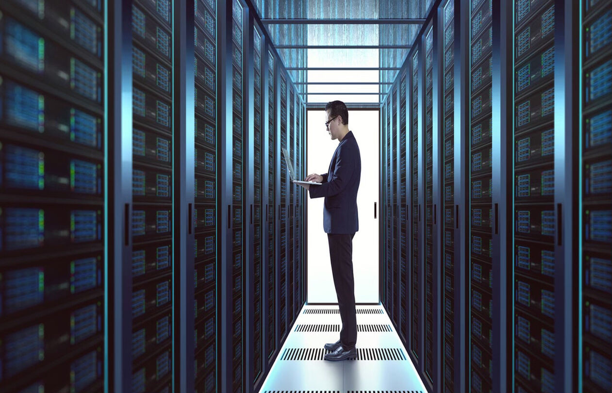 A person in a suit stands in a server room, using a laptop surrounded by rows of server racks.